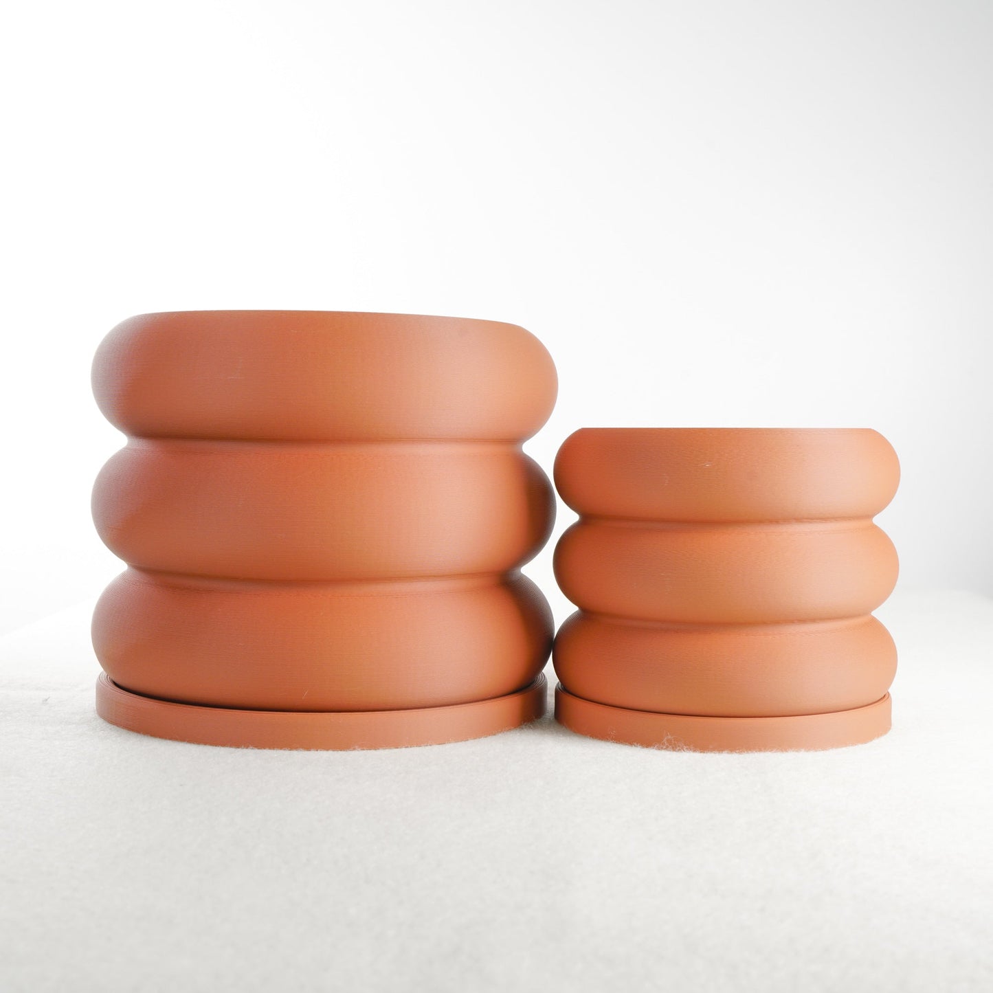 Two terracotta-colored Bubble Planters resembling whimsical stacks of donuts with rounded, bubble-like contours, featuring excellent drainage and matching trays. Ideal for plant lovers and home decor enthusiasts, these planters are available in multiple sizes and colors, adding a touch of modern minimalism to any space.